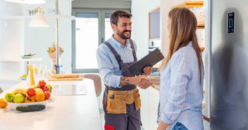 Contractor wearing tool belt holding clipboard smiling shaking hand of woman in kitchen