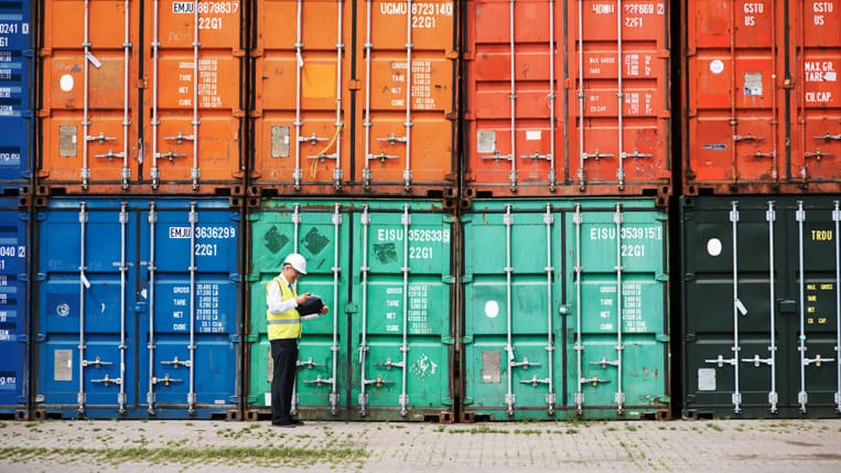 A worker in a hard hat and high visibility vest stands in front of stacked, multicolored shipping containers.