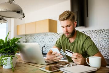 Young bearded man wearing green t-shirt sitting in the kitchen at home, holding smart phone and credit card in hands, shopping online.