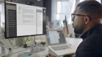  black man’s face looking into a computer screen in an open plan working office. Type is being added to the screen by an Artificial intelligence, AI, chatbot.