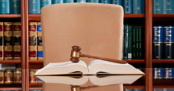 View of law office desk with gavel sitting on top of open book and bookshelf behind large chair.