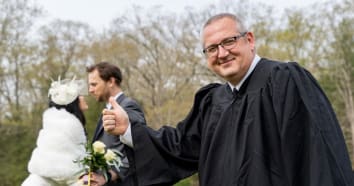 Wedding officiant in a robe with thumbs up and newly married couple in background about to kiss