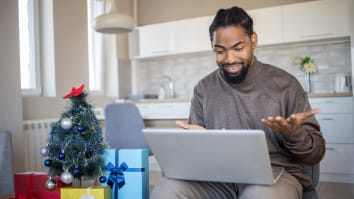 A young man is raising his hands in confusion while looking at something on his laptop. He is seated next to a small Christmas tree surrounded by wrapped presents.