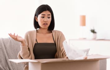 Wrong Item Concept. Dissatisfied unhappy asian woman sitting on sofa with cardboard box, received parcel with wrong or damaged staff, spreading hands in disappointment, feeling upset with bad purchase (Wrong Item Concept. Dissatisfied unhappy asian wo