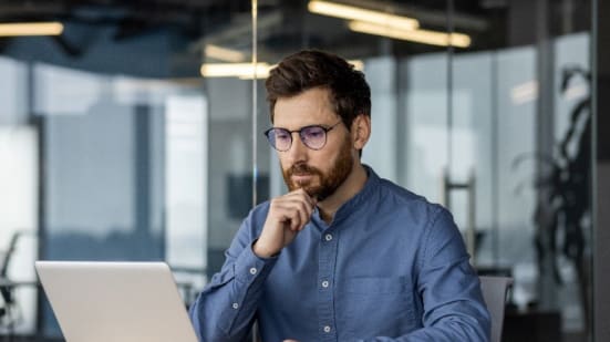 Man with glasses and beard sitting at a desk in a modern office, looking thoughtfully at a laptop screen with his hand resting on his chin. A smartphone and notepad are on the desk beside him