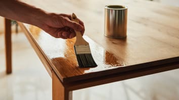 A close-up on someone's hand as they oil a wooden table with a paintbrush.