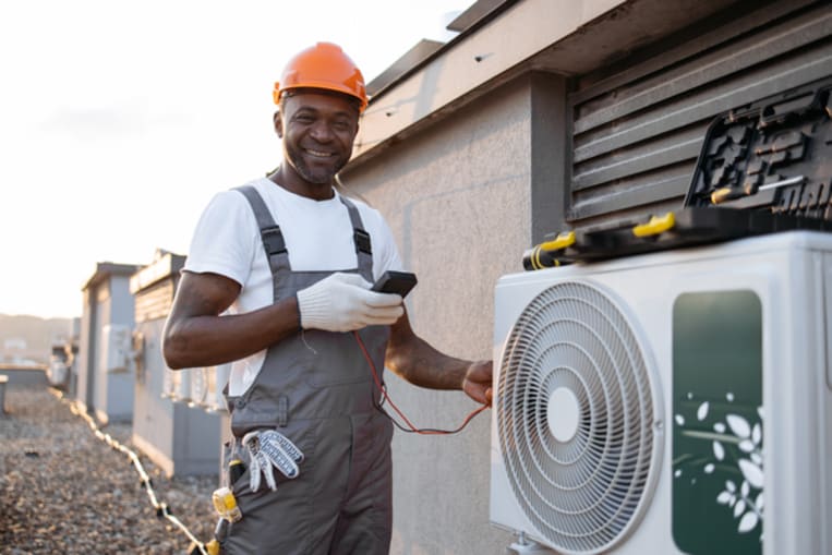 Focused african american specialist using voltage tester while installing modern cooling system on rooftop of residential building. Manager measuring current strength in hanging air conditioner.