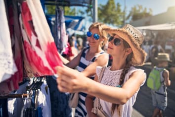 two women wearing sunglasses are smiling and surveying an article of clothing they are considering buying at an outdoor art fair or festival