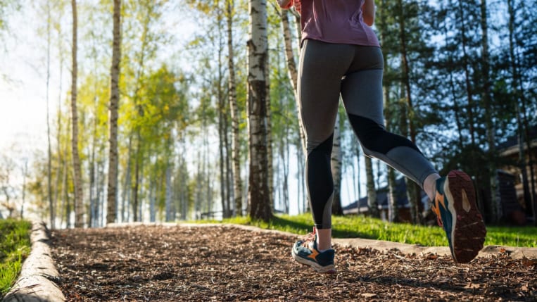 A person is jogging away from the camera down a forested trail, wearing athletic leggings and running shoes.