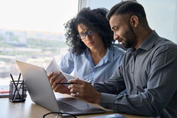 Multiethnic male indian mentor and female African American intern sitting at desk with laptop doing paperwork together discussing project financial report. Corporate business collaboration concept.