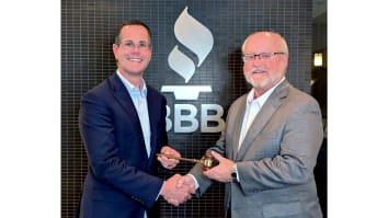 Two men in business suits are shaking hands in front of a wall displaying the BBB Torch logo. One man is handing a gavel to the other.