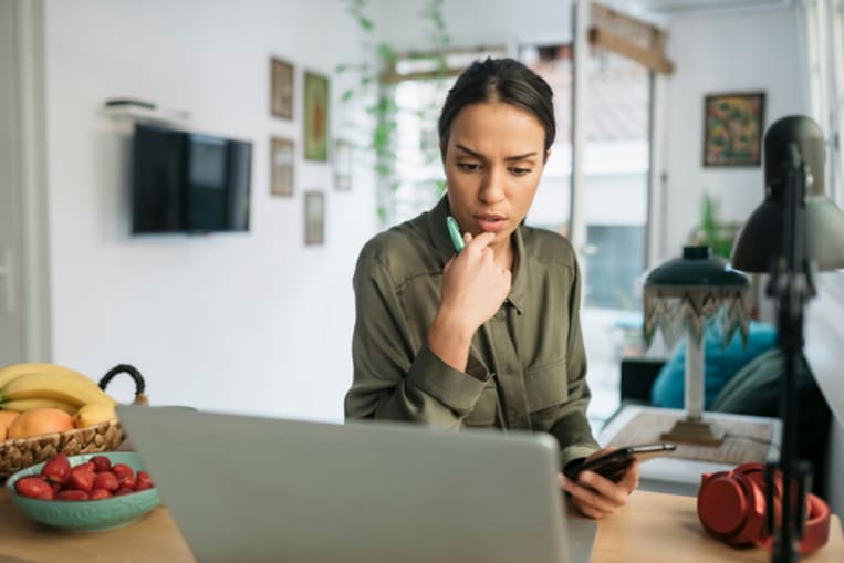 Unemployed beautiful young woman looking for a job online from home using a laptop and her smart phone, trying to decide what to do