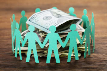Image cutouts of individuals with teal paper that are holding hands and surrounding a stack of hundred dollar bills. 