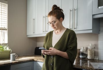 Young redheaded woman leaning against a counter in her kitchen in the morning and sending a text message on her mobile phone