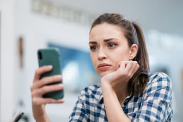 Close up of a Caucasian young woman sitting at home, reading some bad news on her smart phone, feeling sad and worried