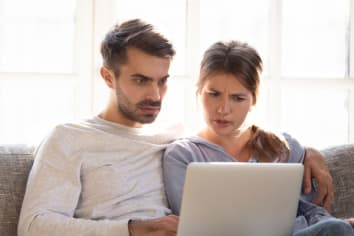 Confused baffled wife and shocked frustrated husband reading online news looking at stuck laptop, bewildered young couple in disbelief perplexed by computer problem email or system failure message