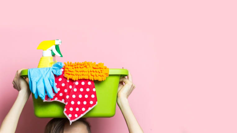 A close-up on a woman standing against a light pink backdrop and holding a tub of cleaning supplies over her head.