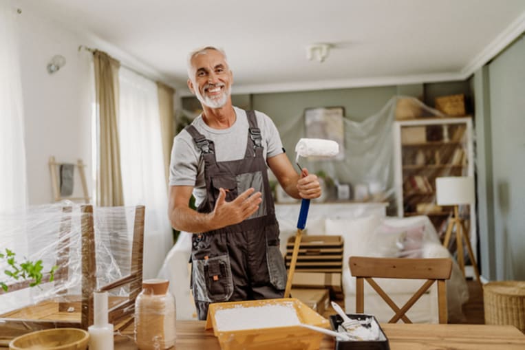 A middle aged male painter wearing a gray shirt and blue overalls looks at the camera smiling while holding a roller.