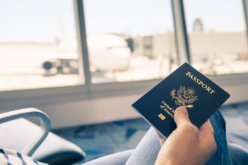 A person holding a U.S. passport while sitting in an airport terminal with a view of an airplane parked at the gate through large windows.