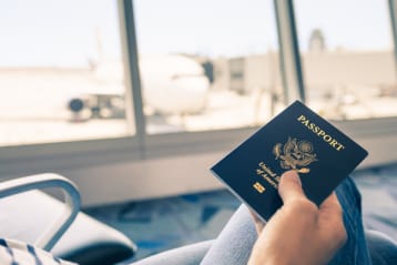 A person holding a U.S. passport while sitting in an airport terminal with a view of an airplane parked at the gate through large windows.