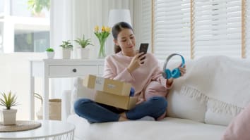 A young woman is sitting on a couch with an open package. She is using her phone to take a picture of a pair of headphones she just removed from the package.