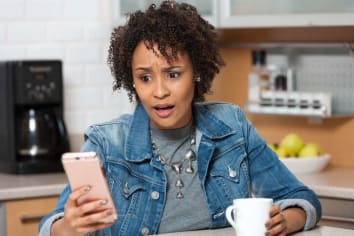 woman holding coffee cup sitting at table looking at phone in shock