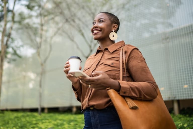 A black woman with short hair smiles looking out to the distance, an office park with trees in the background