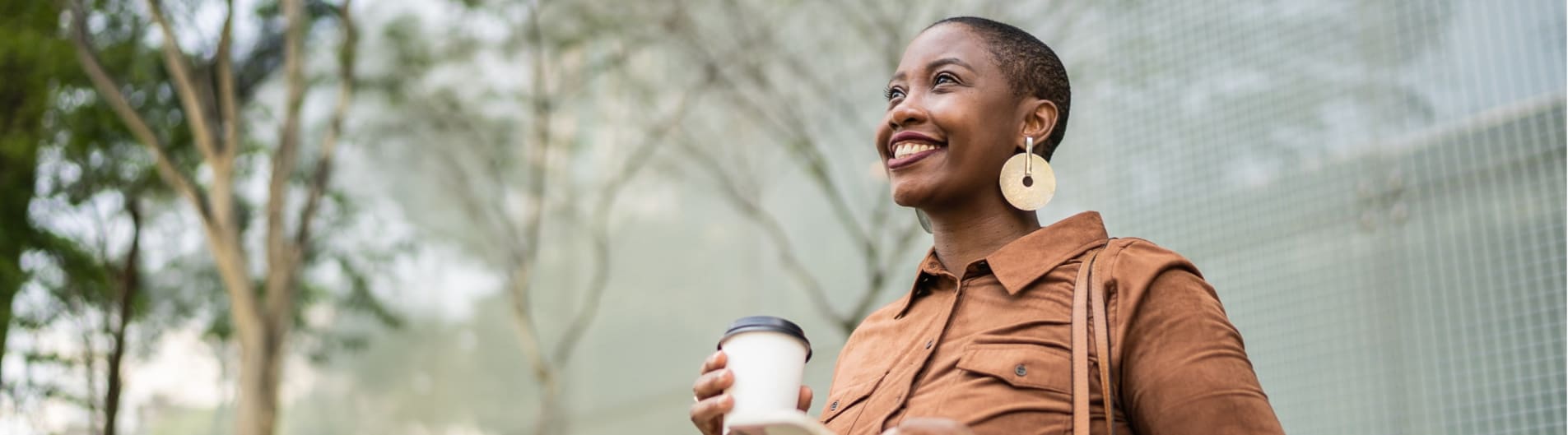 A black woman with short hair smiles looking out to the distance, an office park with trees in the background