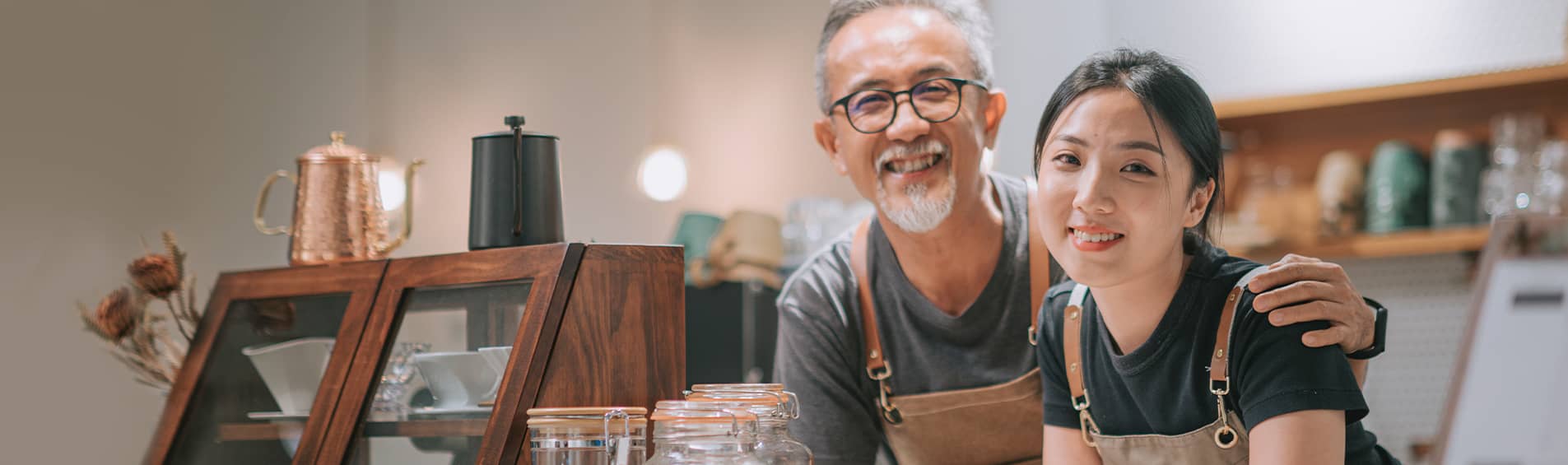A young woman and an older man, both of Asian descent, smile from behind the counter of a cafe. The shop looks modern, and the counter is decorated with goose neck kettles and pourover coffee drippers.