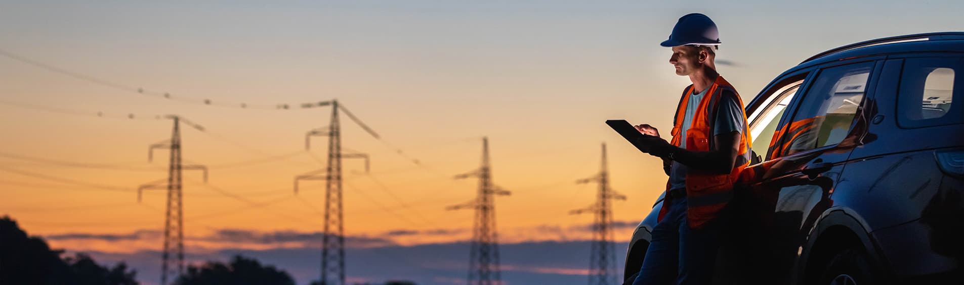 A construction project lead leans against a car, apparently overseeing a project that is out of the shot. In the background, a vast, expansive sunset.