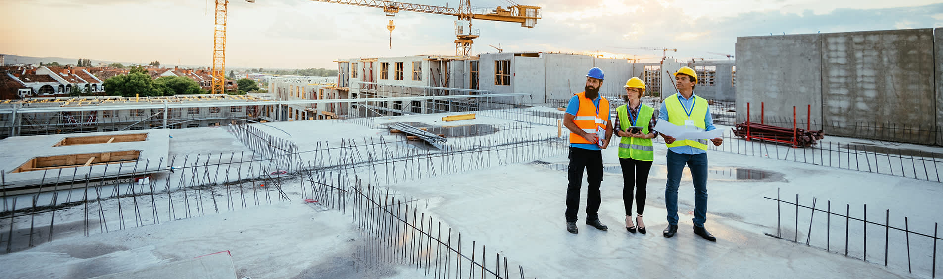 Three construction workers of varied age, race, and gender stand atop an unfinished rooftop construction site, looking towards their work.