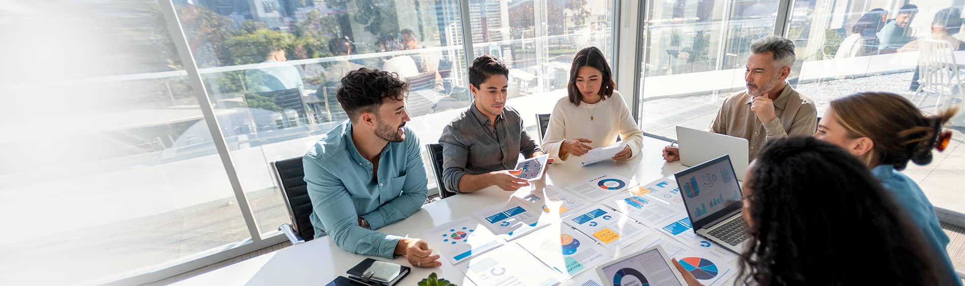 A group of young professionals reviewing sheets of graphs and charts. At the head of the table sits an older man overseeing the meeting.