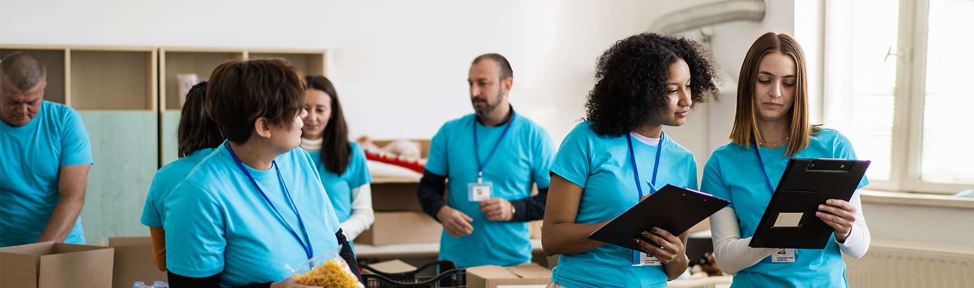 A group of volunteers. In the foreground, a diverse group of women check notes on a clipboard. In the background, others work on assembling boxes.