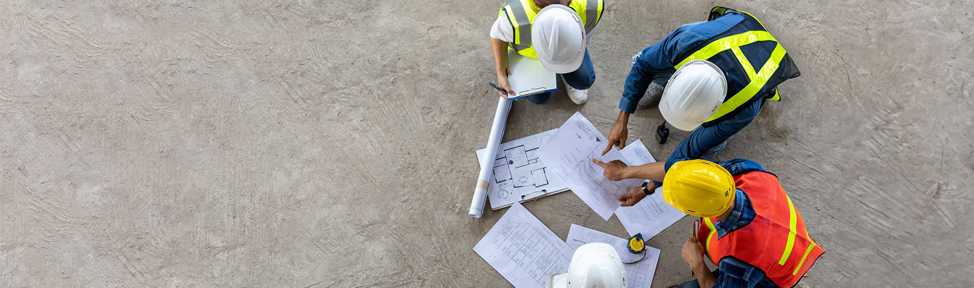 Four construction workers in hard hats and fluorescent vests stand over architectural plans, pointing and writing.