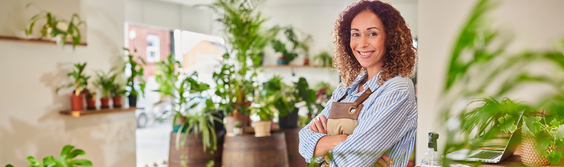 A tan skinned woman with curly hair wearing coveralls smiles in a room full of plants for sale.